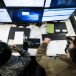 two people sitting at a desk with multiple computer screens