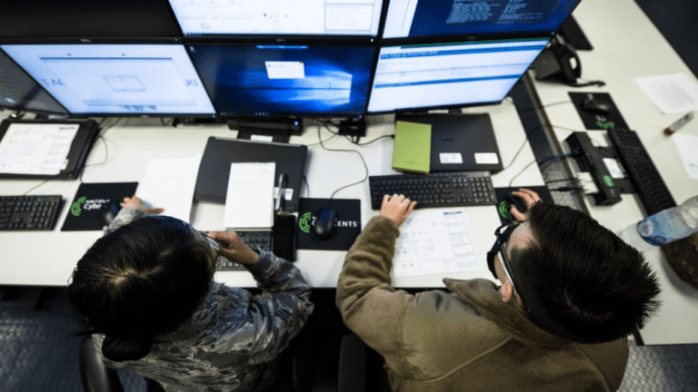 two people sitting at a desk with multiple computer screens