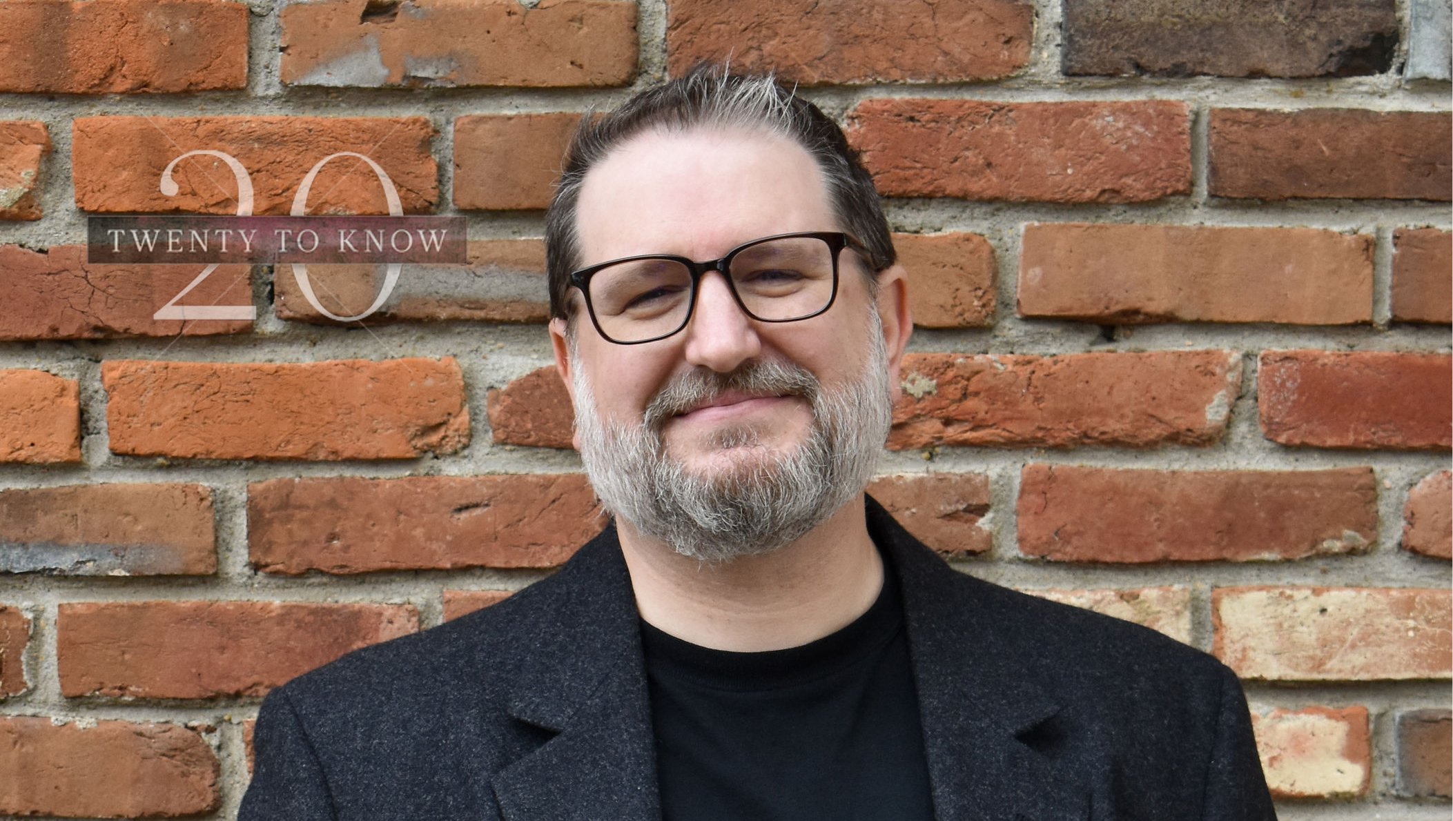 a man with glasses and a beard standing in front of a brick wall